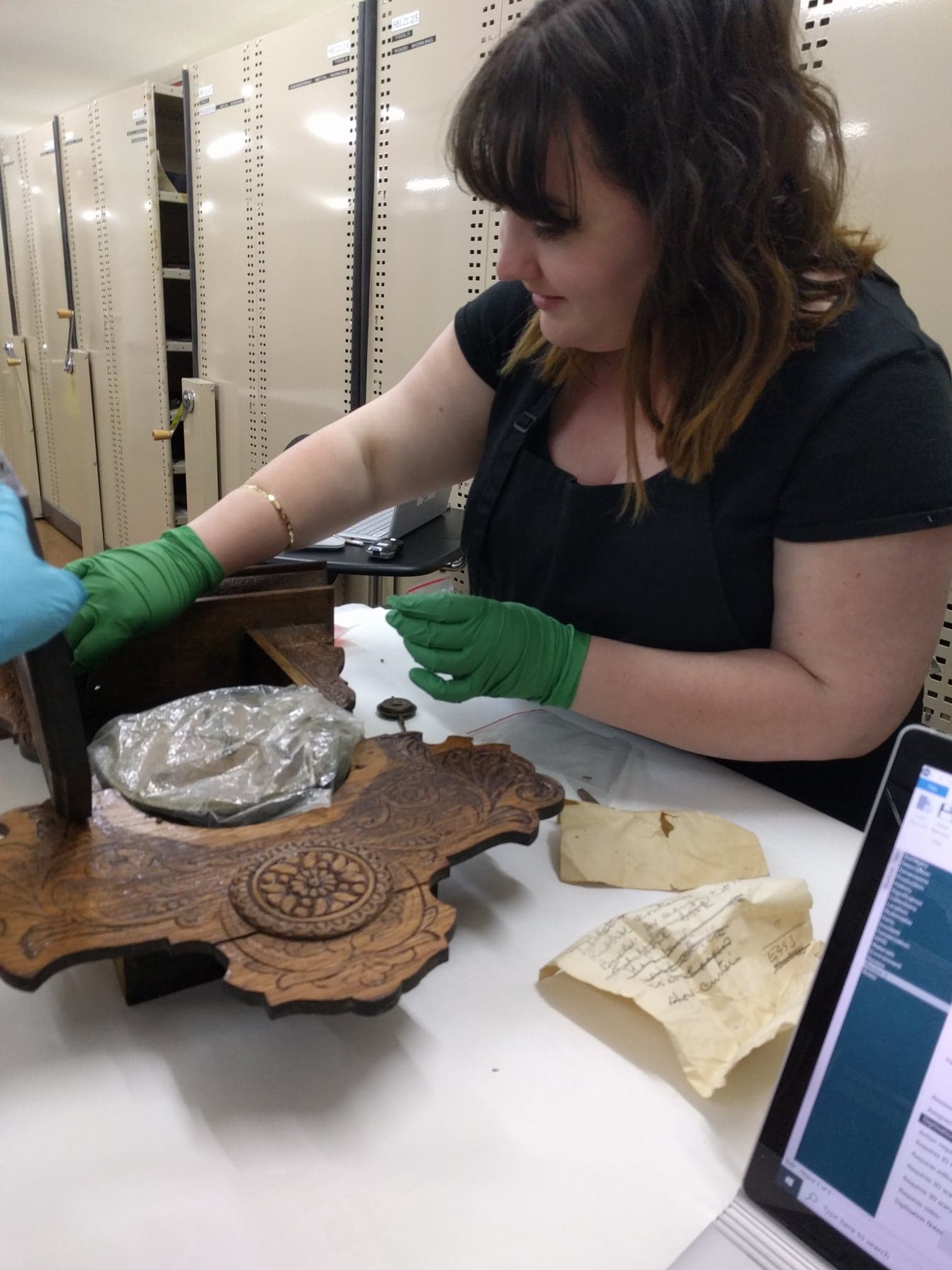 A woman wearing latex gloves in the process of cleaning a carved, wooden collection item. There is a dramatic difference between the cleaned and uncleaned areas, with a dark coating of dust having been removed. Behind her is a row of compactus bays.
